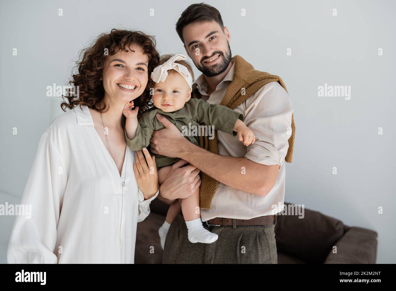 hombre barbudo positivo sosteniendo en brazos a la hija del bebé en la diadema cerca de esposa rizada, imagen de archivo Foto de stock