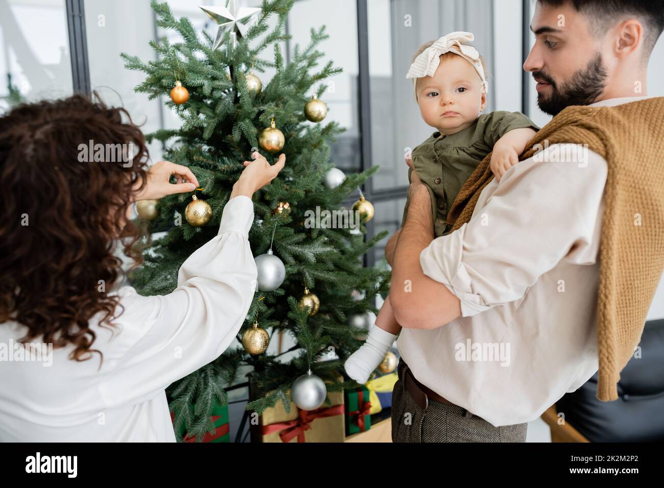 mujer rizada que adorna el árbol de navidad cerca del marido barbudo y la hija infantil, imagen de archivo Foto de stock