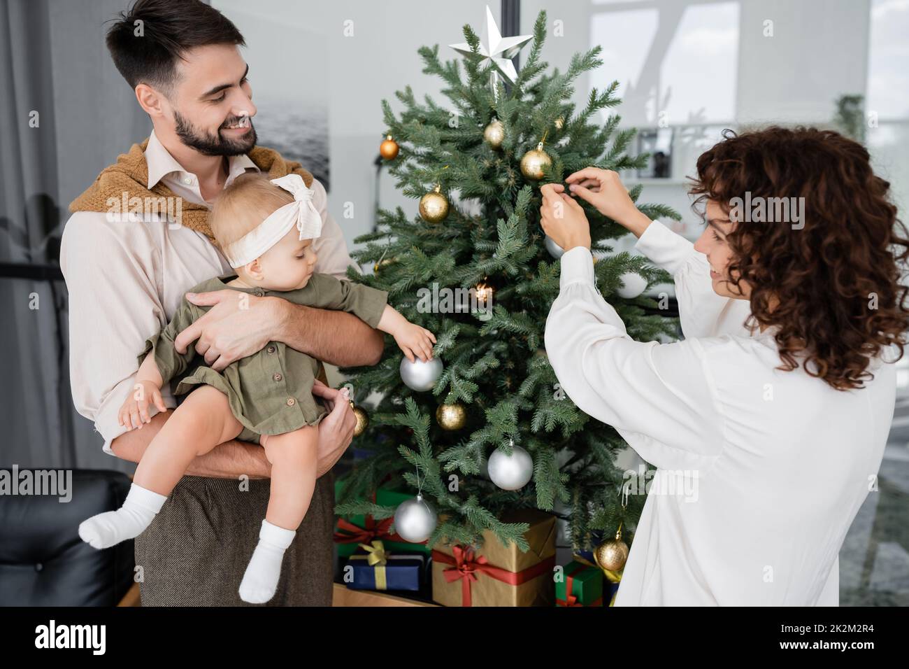 mujer feliz que adorna el árbol de navidad cerca del marido barbudo y la hija del bebé, imagen de la acción Foto de stock
