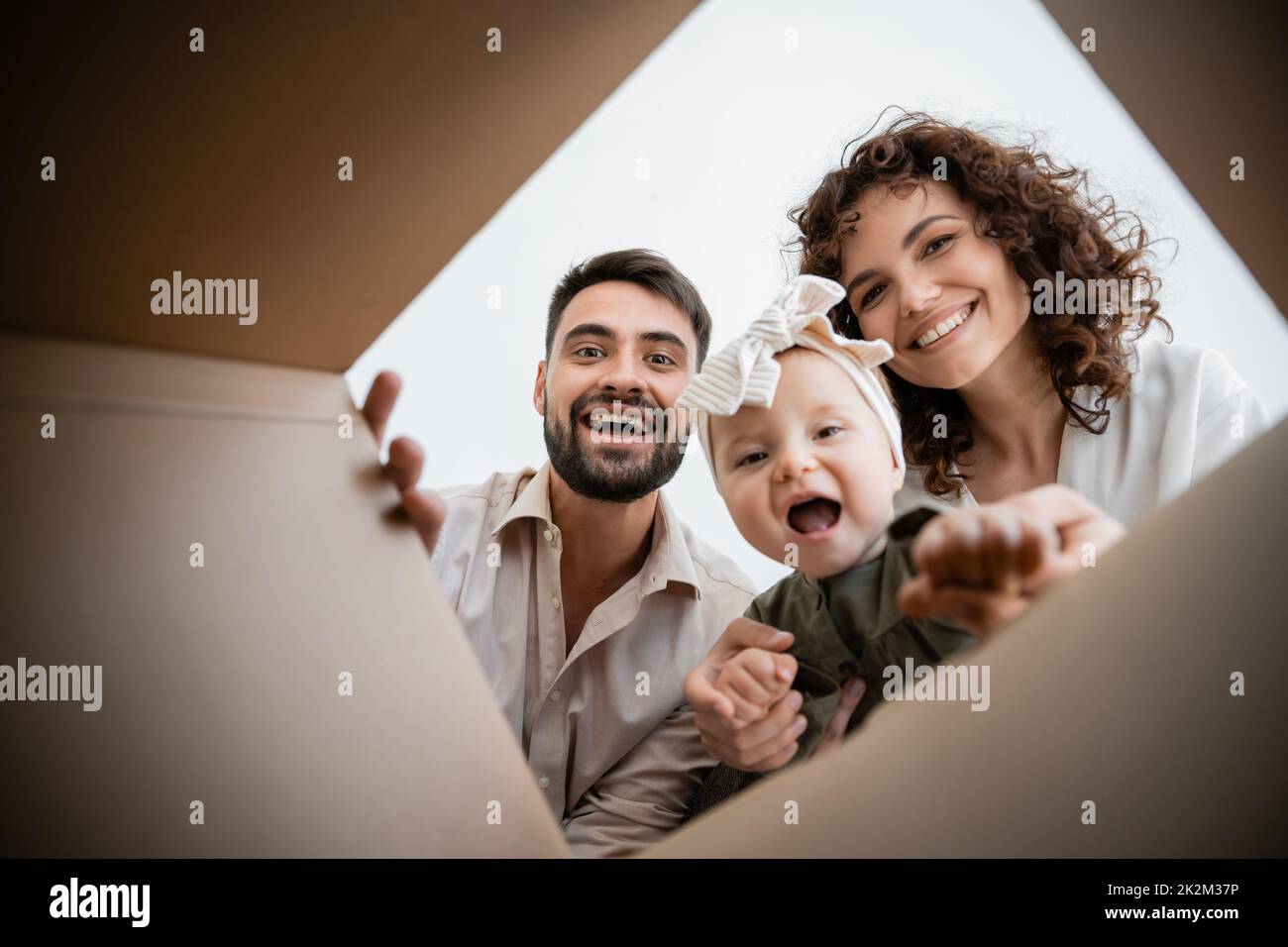 vista inferior de los padres felices y la niña infante emocionada mirando dentro de la caja de cartón, imagen de stock Foto de stock