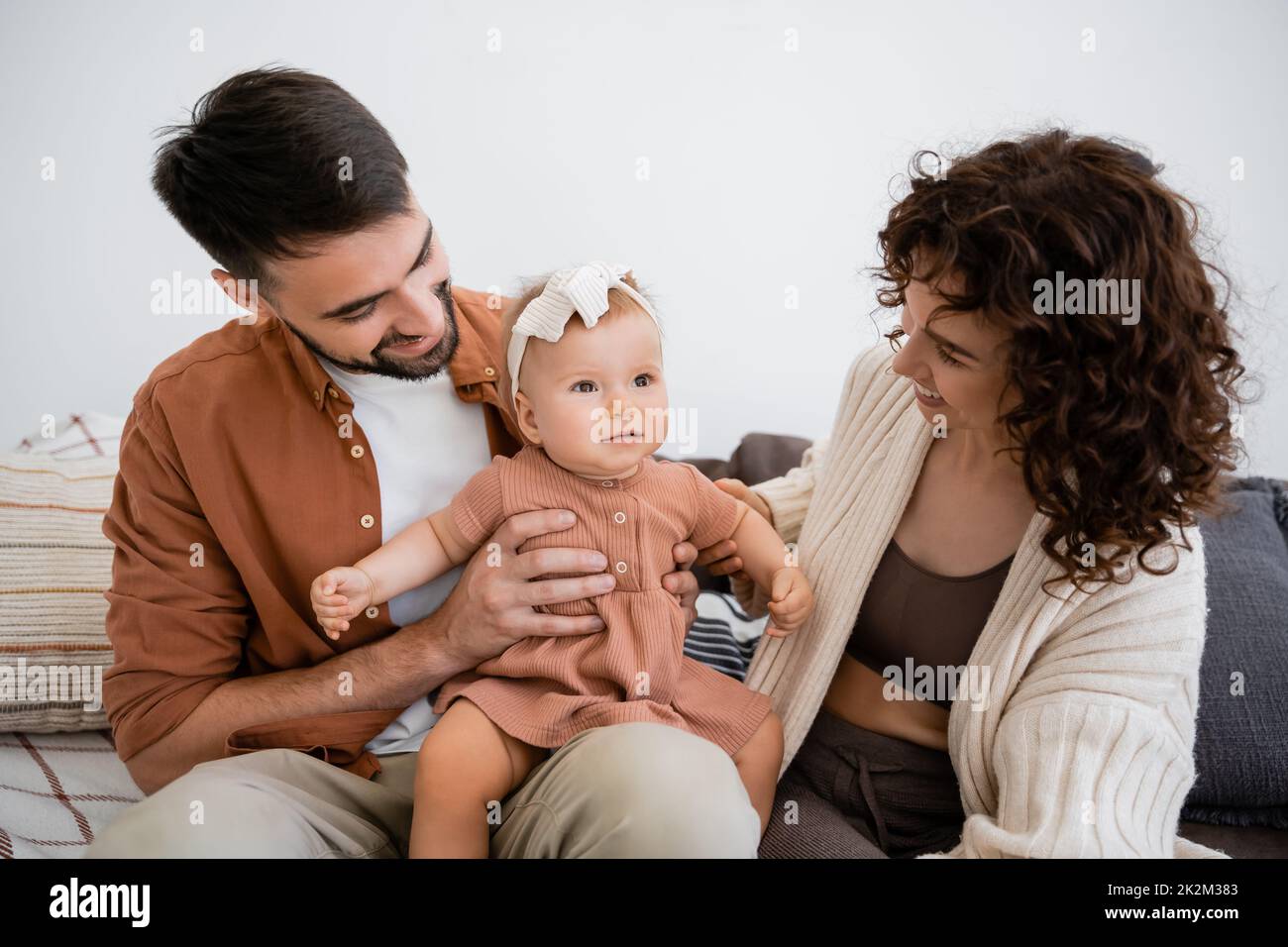 padre alegre sosteniendo feliz hija infantil cerca de la esposa rizada sentada en el sofá, imagen de archivo Foto de stock