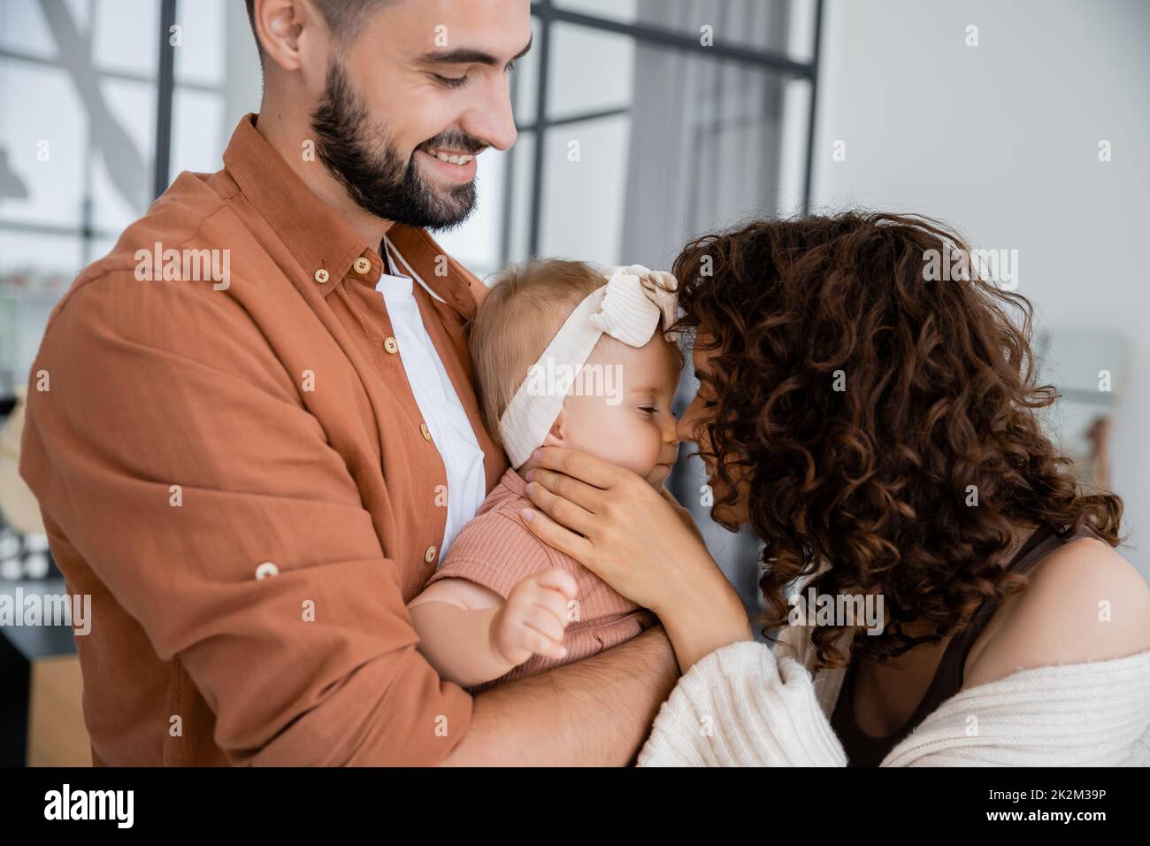 rizado madre tocando la nariz de bebé hija en la diadema cerca de marido alegre en casa, imagen de archivo Foto de stock