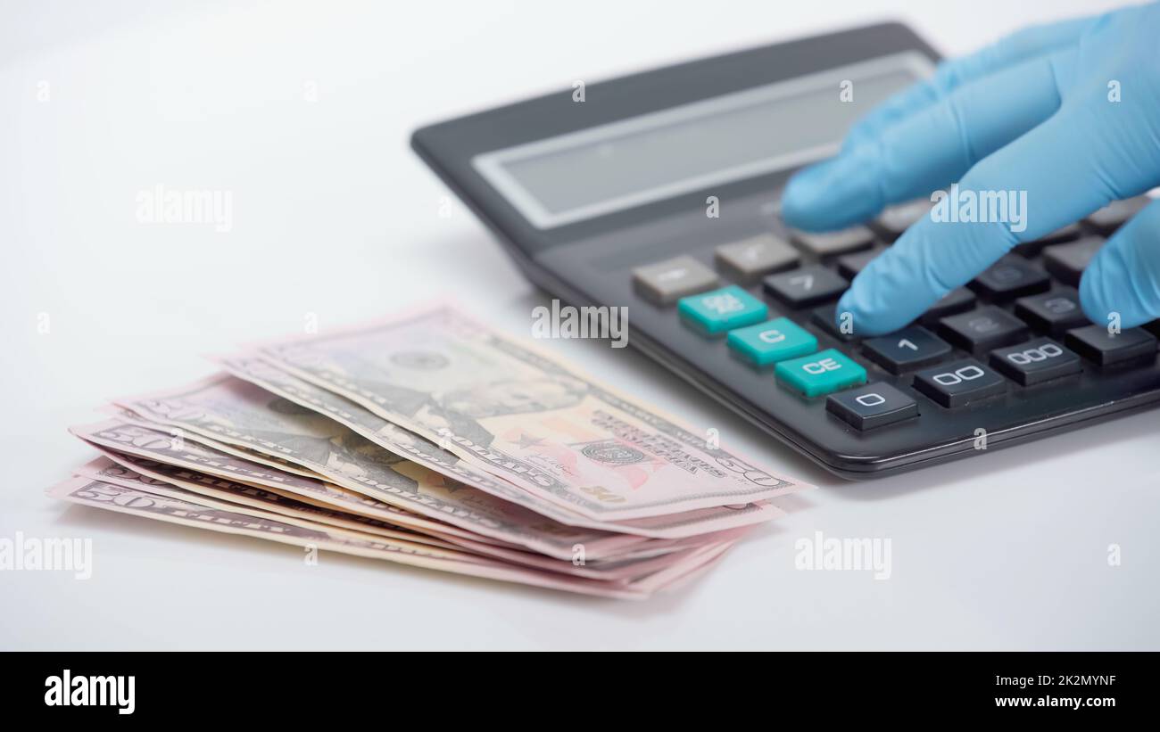 vista recortada de una persona en un guante de látex usando una calculadora cerca de dólares sobre fondo blanco Foto de stock