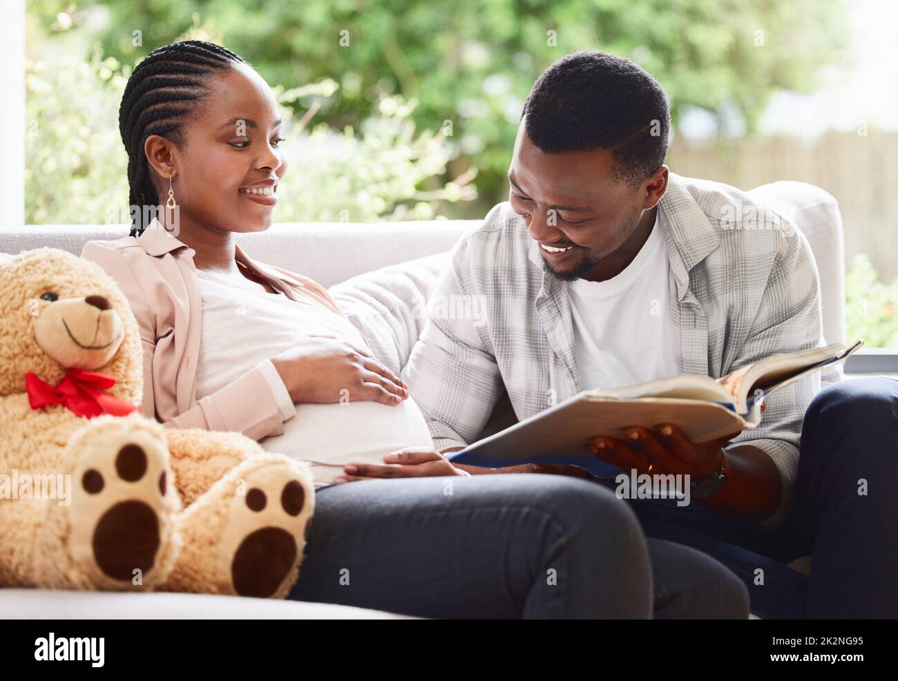 Le encanta leer a su bebé nonato. Foto recortada de un hombre joven guapo leyendo un libro a las mujeres embarazadas vientre mientras se sienta en el sofá en casa. Foto de stock