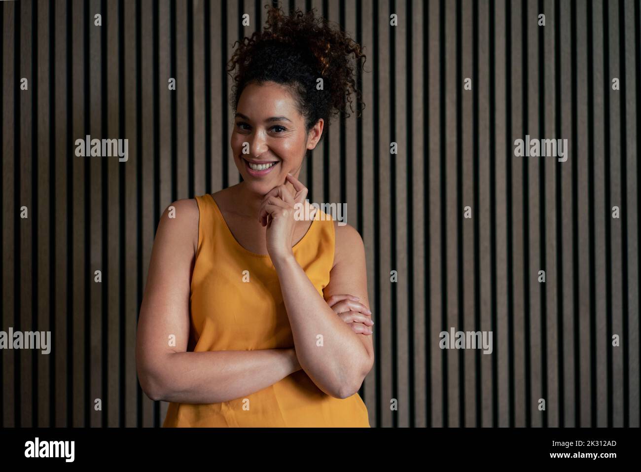 Mujer sonriente con la mano en la barbilla delante de la pared a rayas marrones Foto de stock