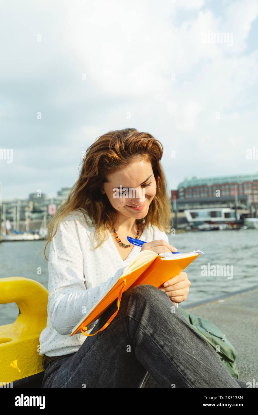 Mujer sonriente con bolígrafo escribiendo en el diario en el paseo marítimo Foto de stock