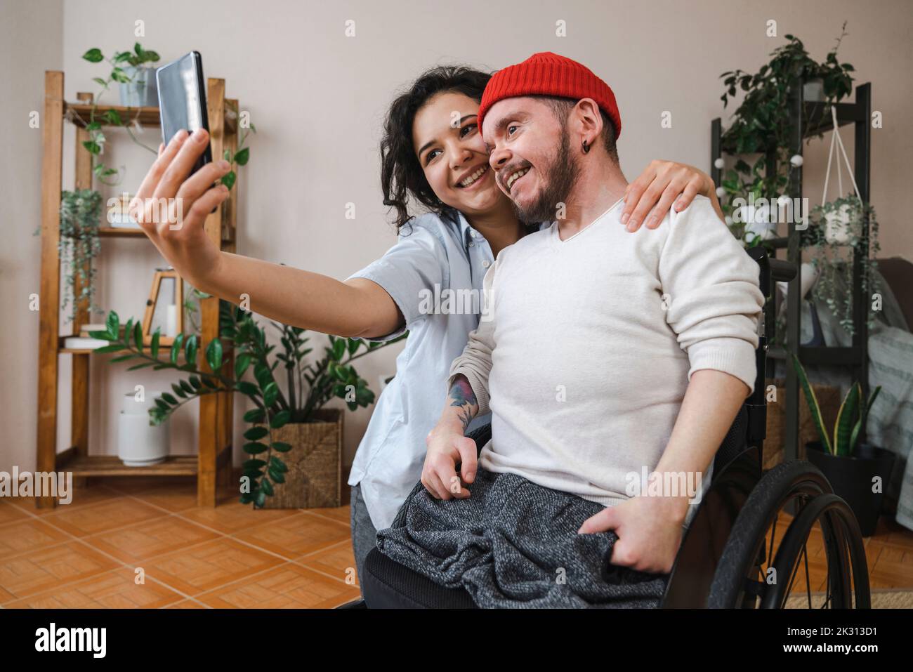 Mujer sonriente con novio en silla de ruedas tomando selfie a través del teléfono móvil en casa Foto de stock