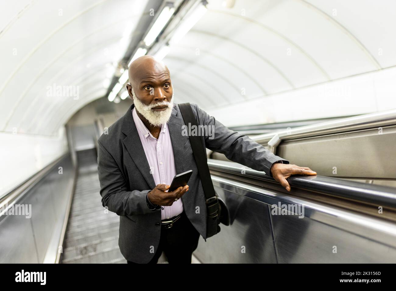 Hombre de negocios de la tercera edad con un teléfono inteligente subiendo por la escalera mecánica Foto de stock