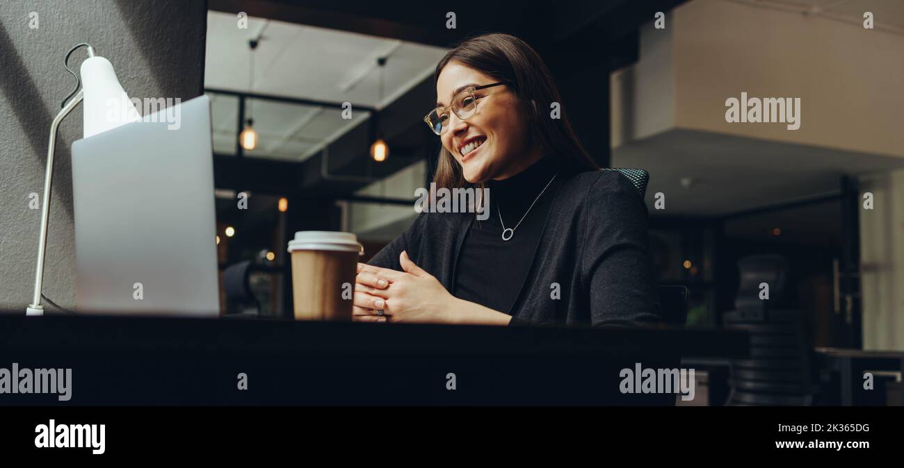 Busineswoman sonriendo alegremente durante una reunión en línea en una oficina moderna del co-trabajo. Empresaria feliz comunicándose con su b Foto de stock