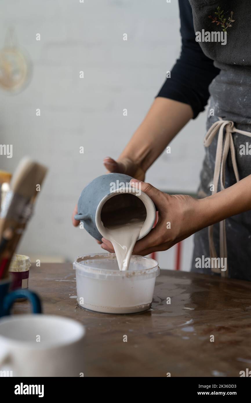 La artesana vierte el líquido de la suciedad del jarrón gris en el tazón de plástico para limpiar el producto para la exposición Foto de stock