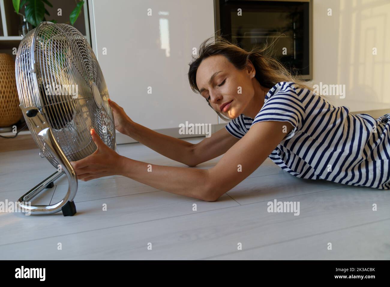 Mujer sudorosa acostada en el suelo con la cabeza hacia el dispositivo sostiene el ventilador e intenta atrapar la corriente de aire frío Foto de stock
