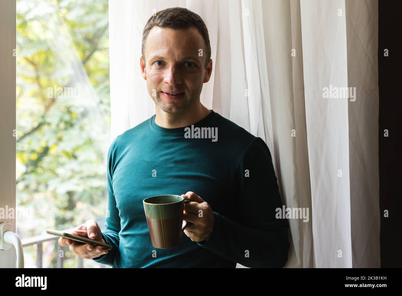 Retrato casero casual de un hombre caucásico adulto joven levemente sonriente con una taza de café y un smartphone en sus manos Foto de stock