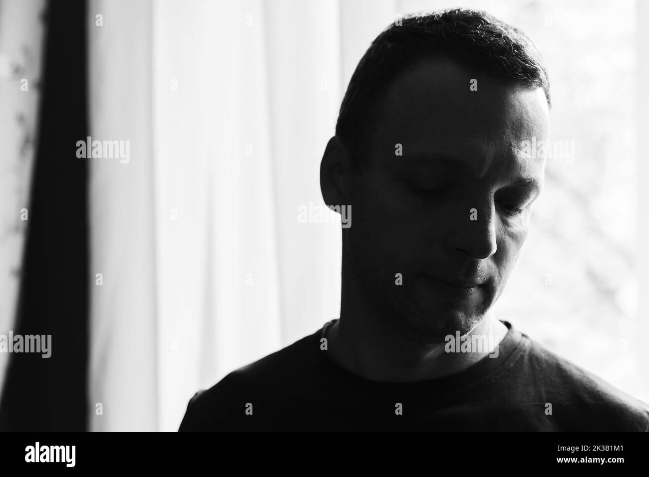 Retrato de estudio oscuro de un hombre caucásico adulto joven cerca de una ventana iluminada brillante, fotografía artística en blanco y negro con enfoque selectivo suave Foto de stock