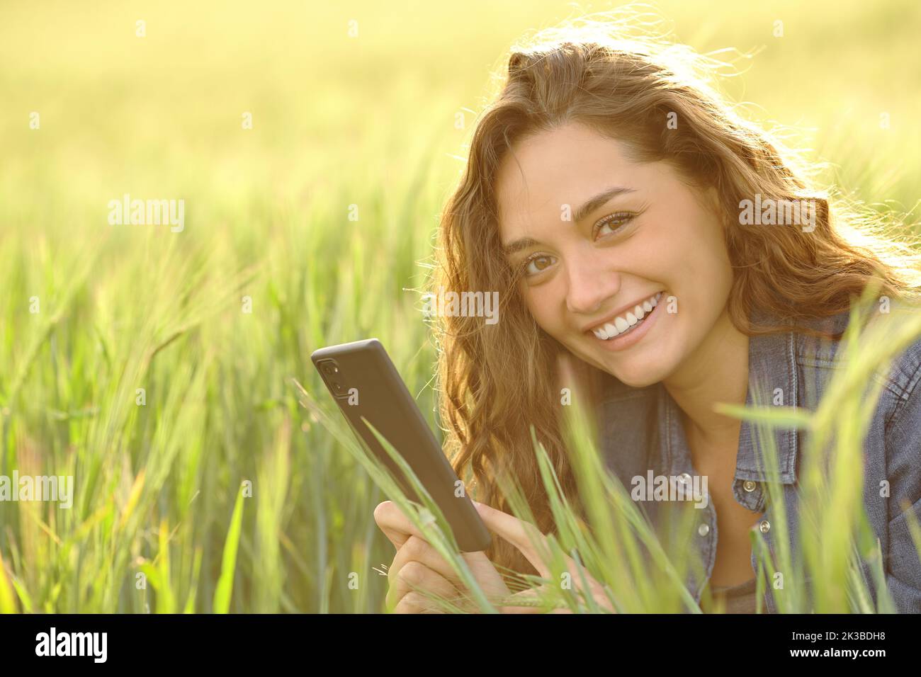 Mujer feliz sosteniendo un teléfono inteligente mirando la cámara en un campo de trigo Foto de stock