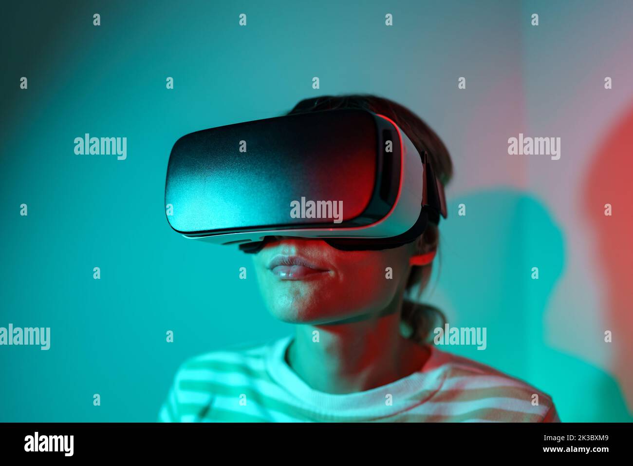 Mujer joven asombrada probando casco moderno de alta tecnología VR, obteniendo experiencia 3D en casa. Luz de neón. Foto de stock