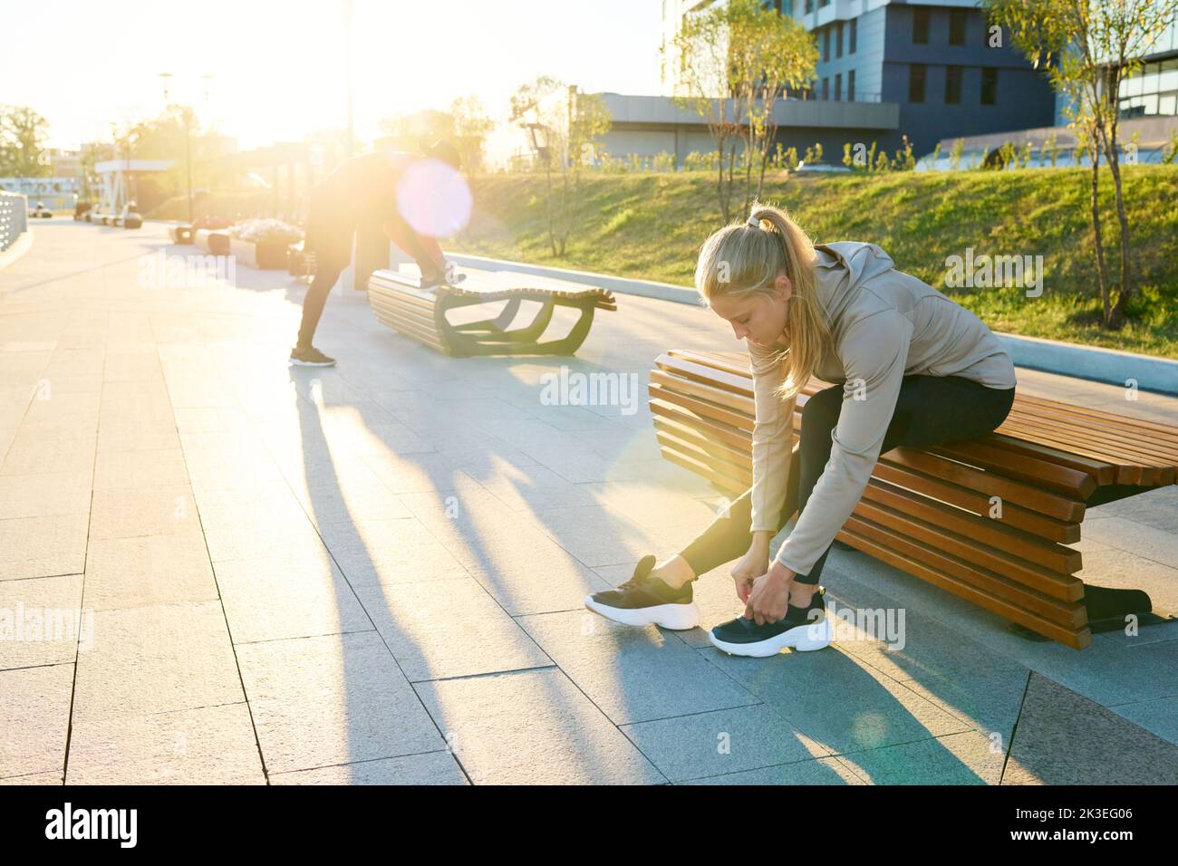 Joven deportista rubia en ropa activa sentada en la banca en un entorno urbano y doblándose sobre las sneakers mientras ataba el zapato Foto de stock