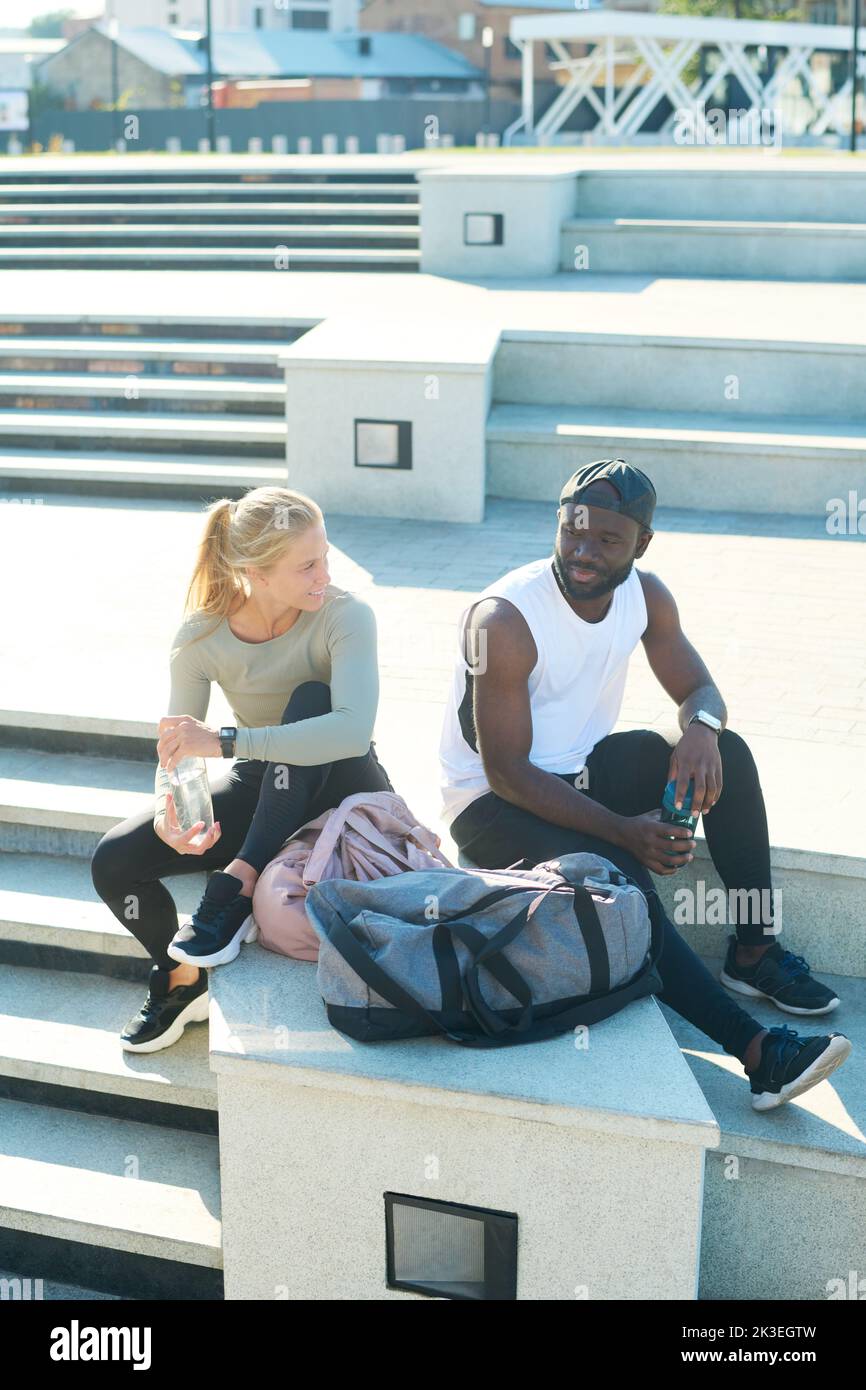 Jóvenes atletas interraciales en ropa deportiva sentados por escaleras al aire libre después de hacer ejercicio o correr y tener algo de agua y charlar Foto de stock