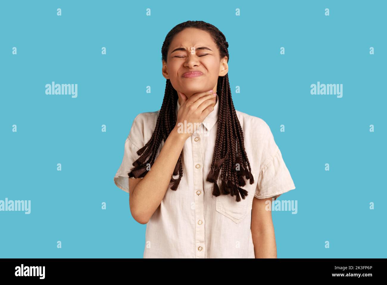 La mujer infeliz con dreadlocks tiene dolor terrible de garganta, sufre de sofocación, toca el cuello, cara fruncida el ceño, difícil de tragar, con camisa blanca. Estudio de interior aislado sobre fondo azul. Foto de stock