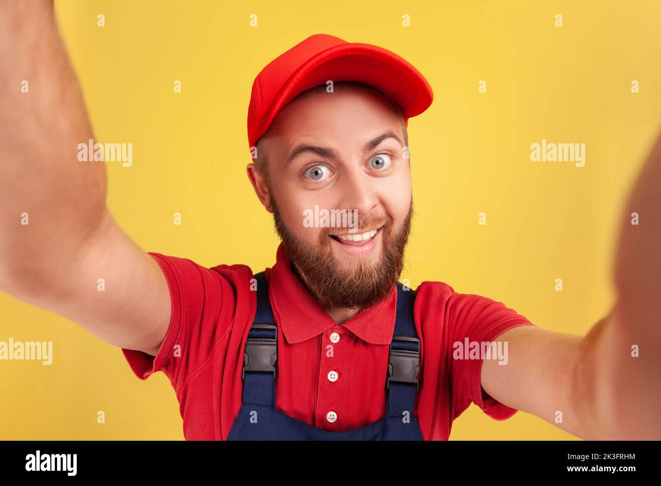 Optimista hombre feliz con el uso de gorra roja y el uniforme azul selfie,  mirando a cámara divertida expresión facial POV, punto de vista de la foto.  Estudio de interior grabado aislado