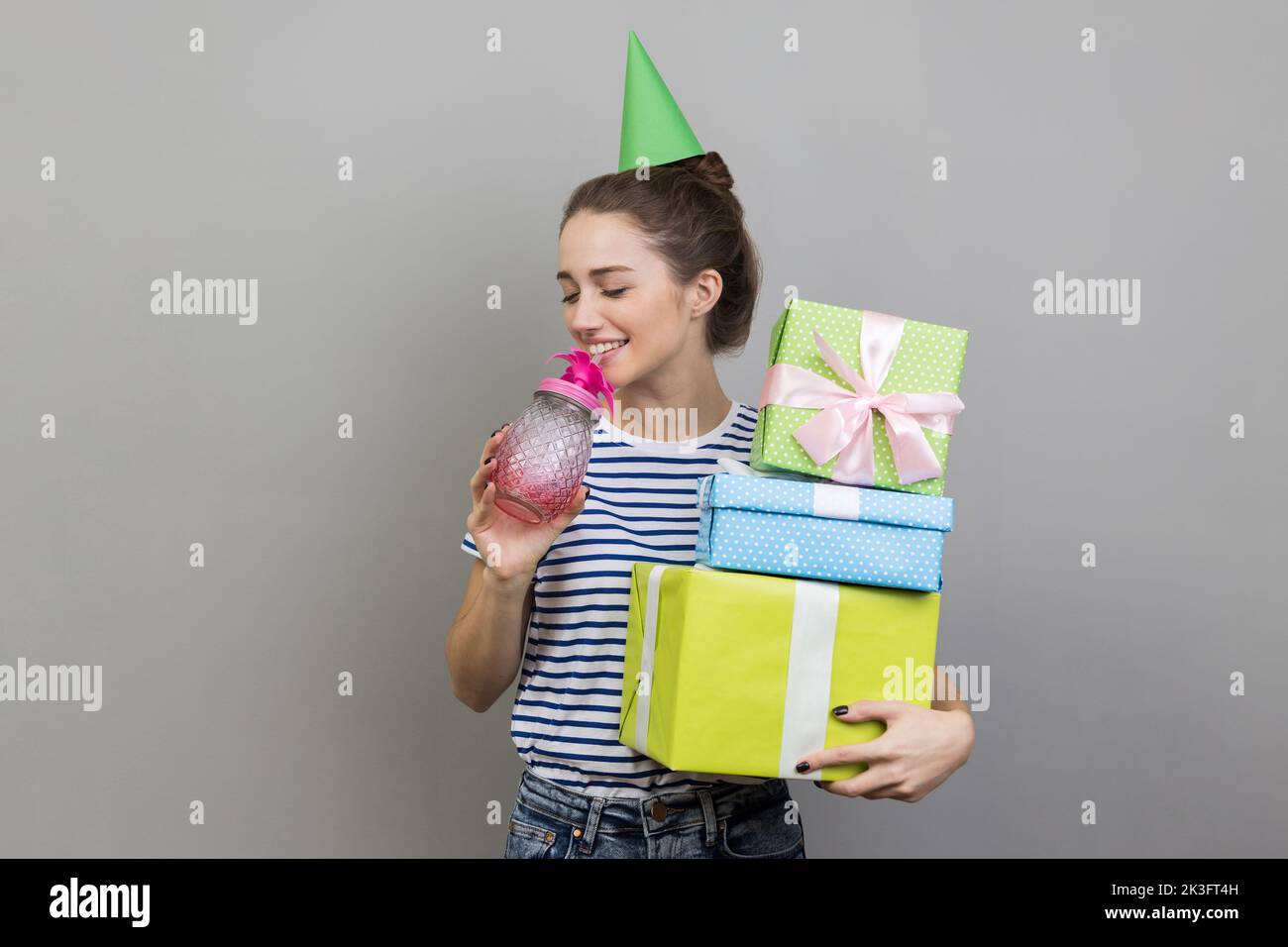 Retrato de una mujer positiva festiva con camiseta a rayas y cono de fiesta, sosteniendo la pila de cajas de regalo, bebiendo cóctel, disfrutando de la fiesta de cumpleaños. Estudio de interior grabado aislado sobre fondo gris. Foto de stock