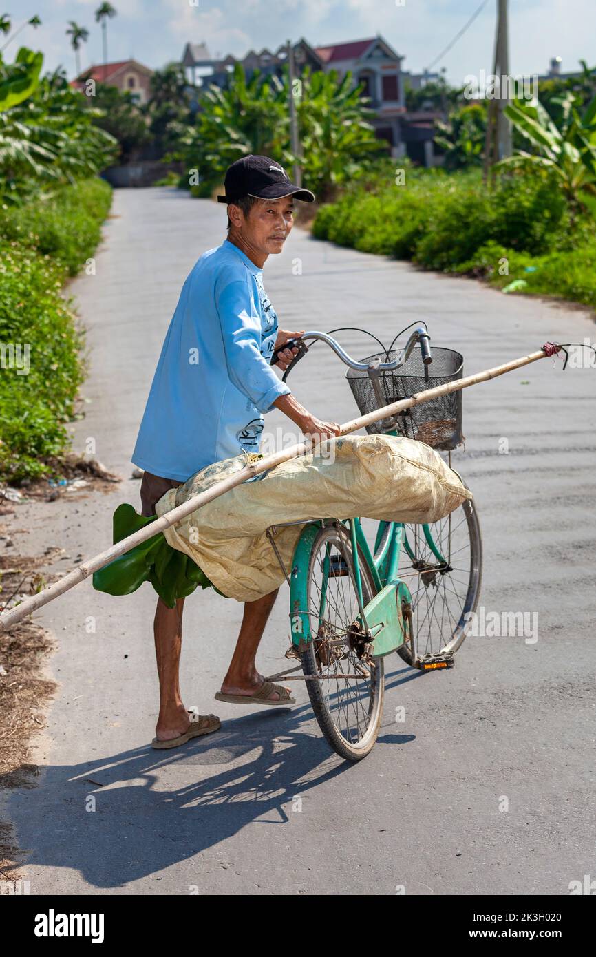Hombre vietnamita llevando un saco en bicicleta en un camino rural pavimentado vacío, Hai Phong rural, Vietnam Foto de stock