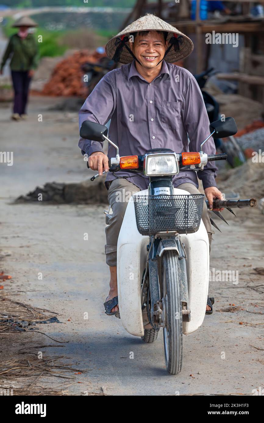 Hombre vietnamita usando sombrero de bambú montando en motocicleta a través del pueblo, rural Hai Phong, Vietnam Foto de stock