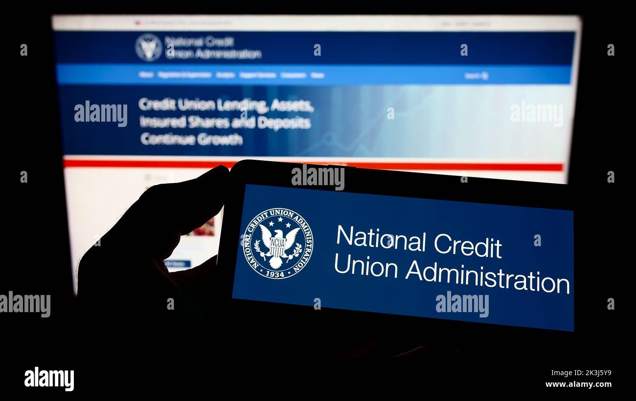 Persona que sostiene un teléfono móvil con el logotipo de la US National Credit Union Administration (NCUA) en la pantalla delante de la página web. Enfoque la pantalla del teléfono. Foto de stock