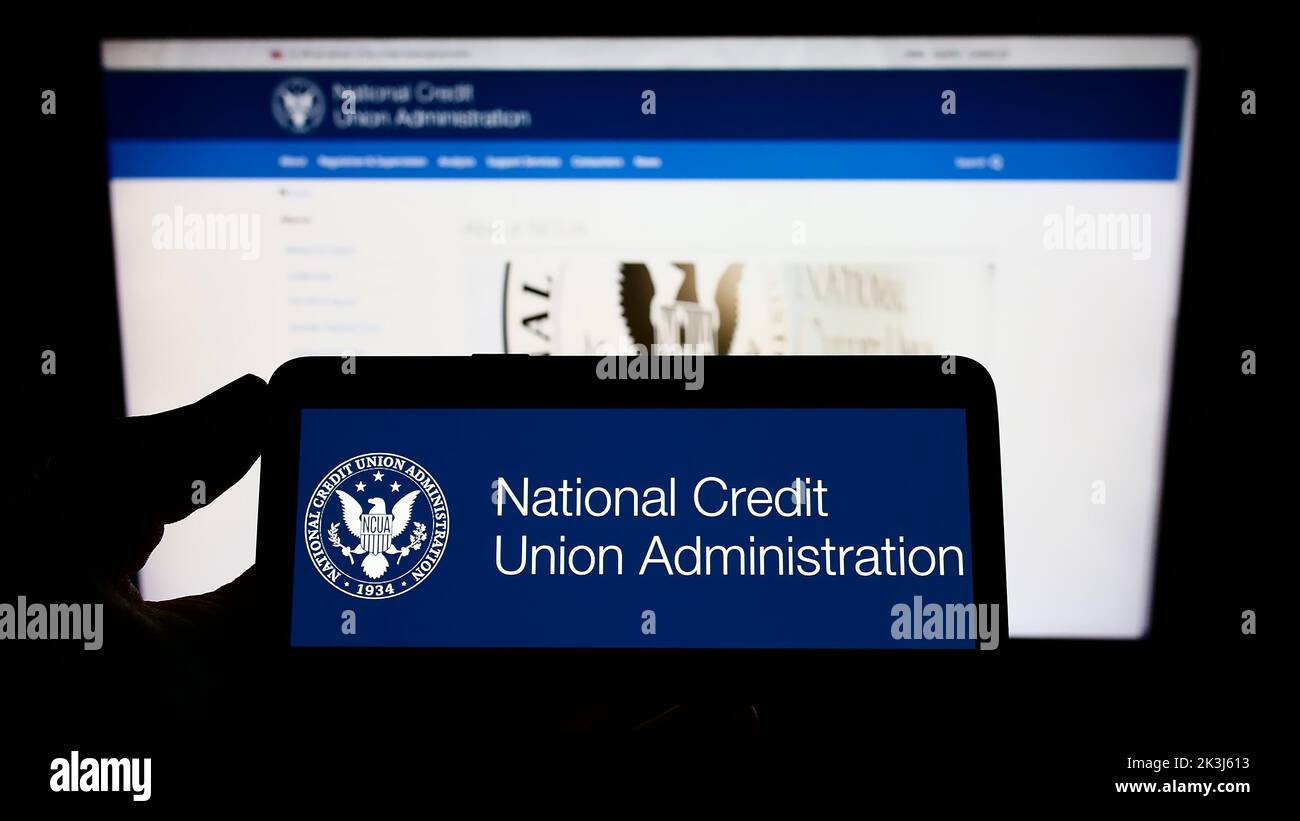 Persona que sostiene un teléfono celular con el logotipo de la American National Credit Union Administration (NCUA) en la pantalla frente a la página web. Enfoque la pantalla del teléfono. Foto de stock
