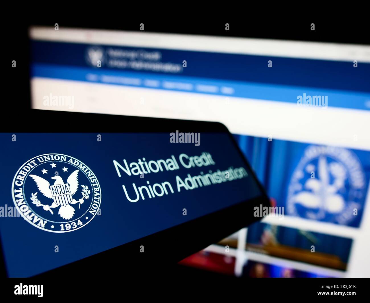 Teléfono celular con el logotipo de la Administración Nacional de Cooperativas de Ahorro y Crédito de los Estados Unidos (NCUA) en la pantalla frente al sitio web. Enfoque en la parte izquierda de la pantalla del teléfono. Foto de stock
