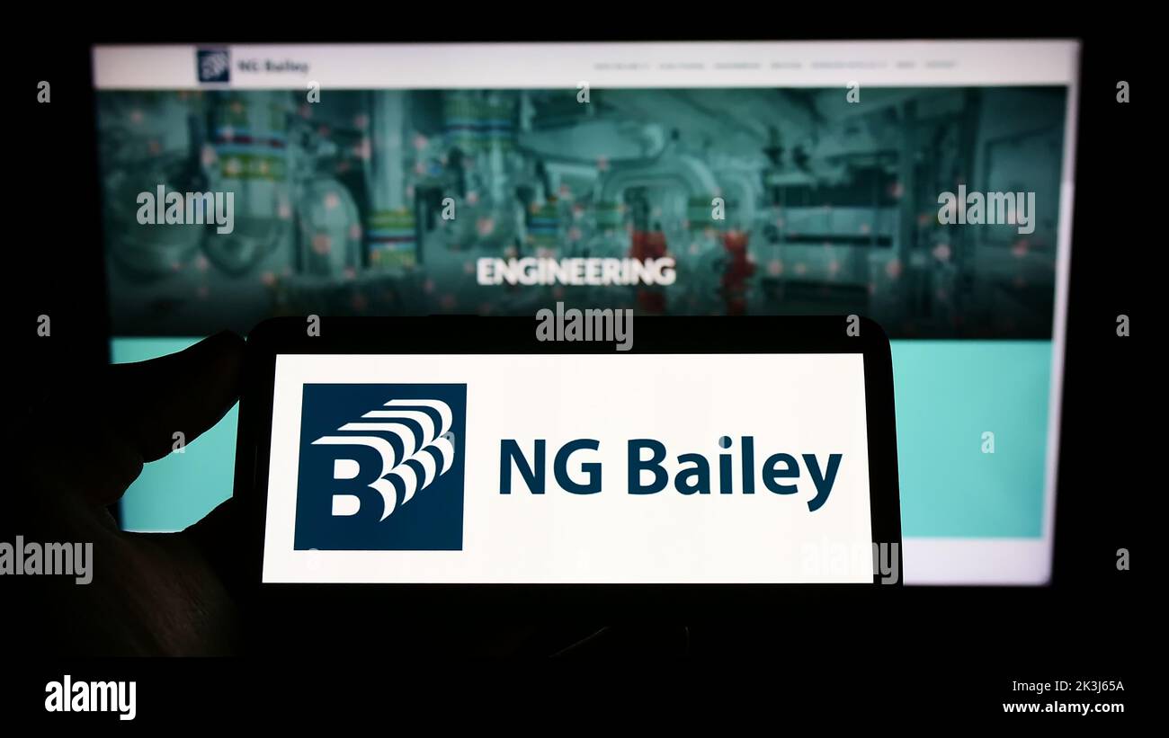 Persona que sostiene el teléfono celular con el logotipo de la empresa de ingeniería NG Bailey Group Limited en la pantalla delante de la página web del negocio. Enfoque la pantalla del teléfono. Foto de stock