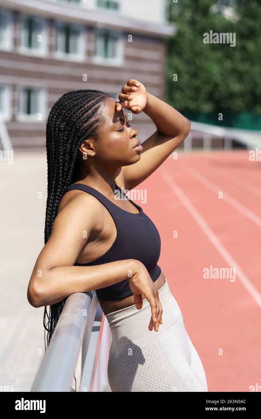 Mujer africana cansada cerrando los ojos con expresión cansada después de correr en la pista roja en un día soleado Foto de stock