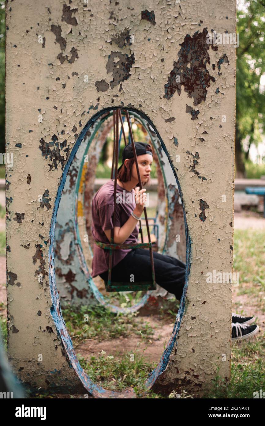 Joven mujer punk sentada en un columpio en el patio de recreo abandonado en verano Foto de stock