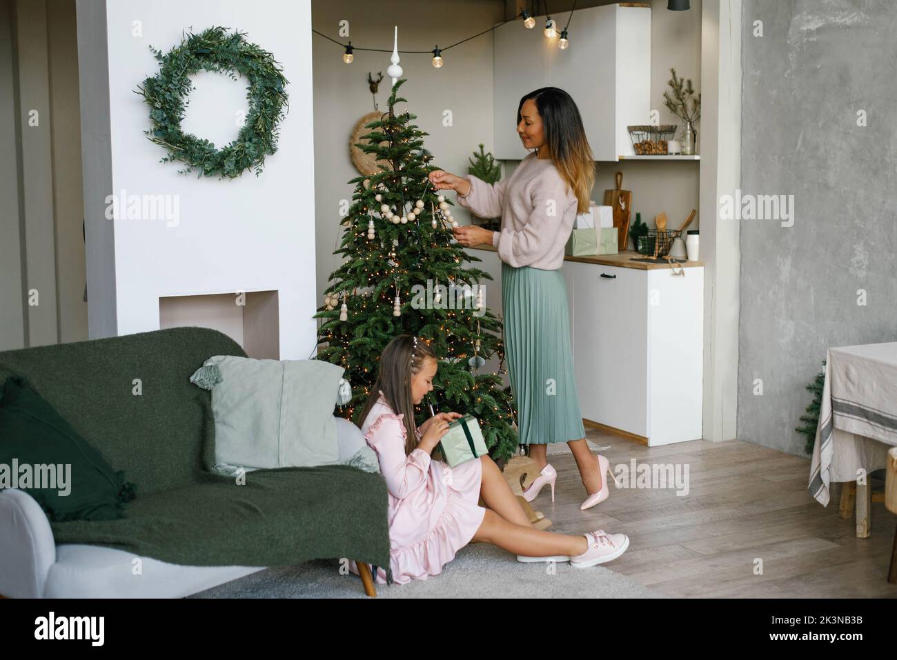 Mamá decora el árbol de Navidad, hija se sienta, abre un regalo de Navidad Foto de stock