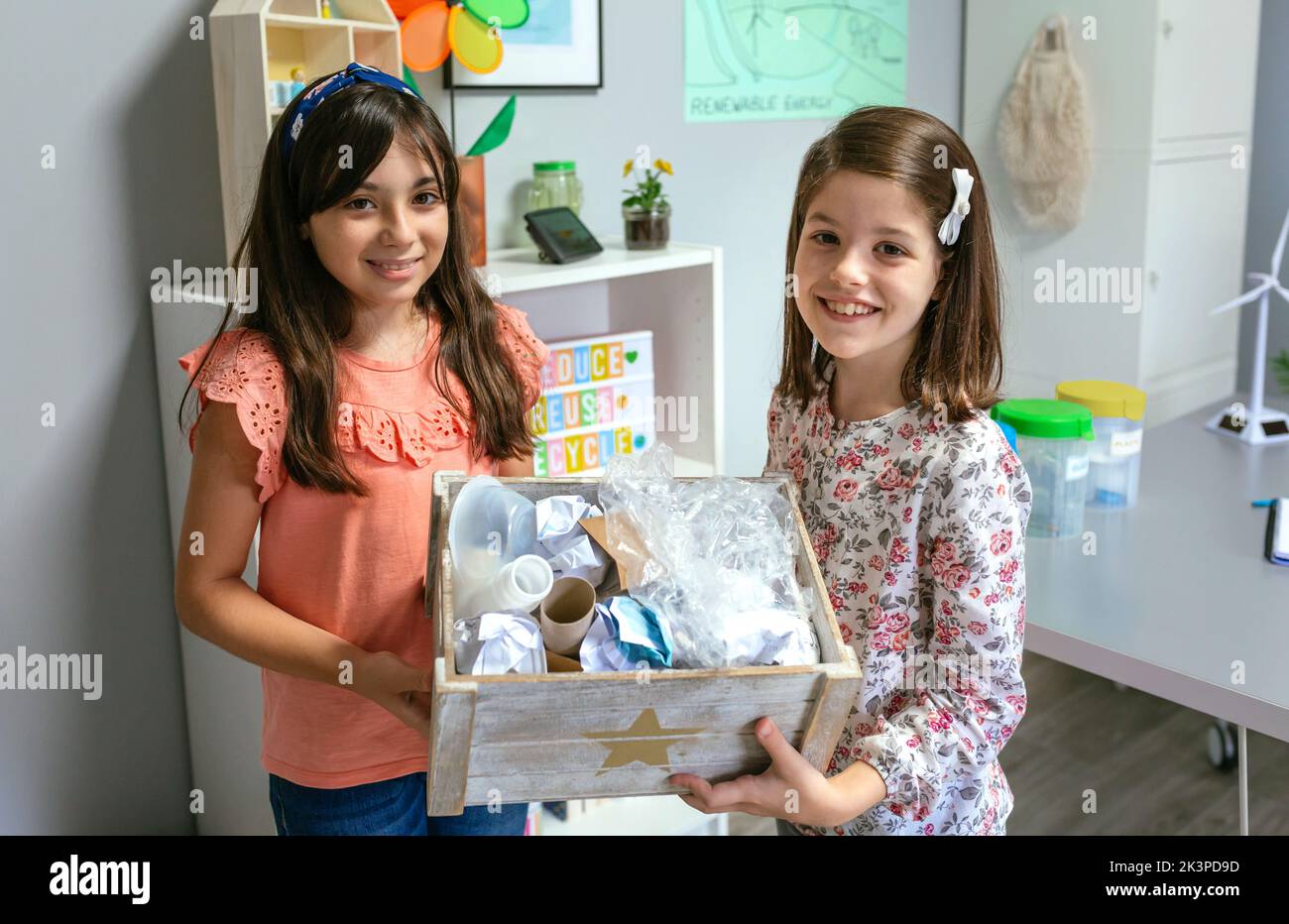 Dos niñas en el aula de ecología mostrando una caja de residuos para reciclar Foto de stock