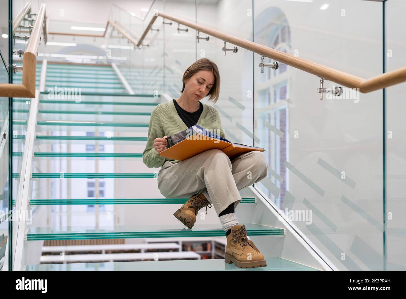 Curiosa lectora se sienta en las escaleras de vidrio moderno paso de lectura de libro favorito sobre la historia del mundo Foto de stock