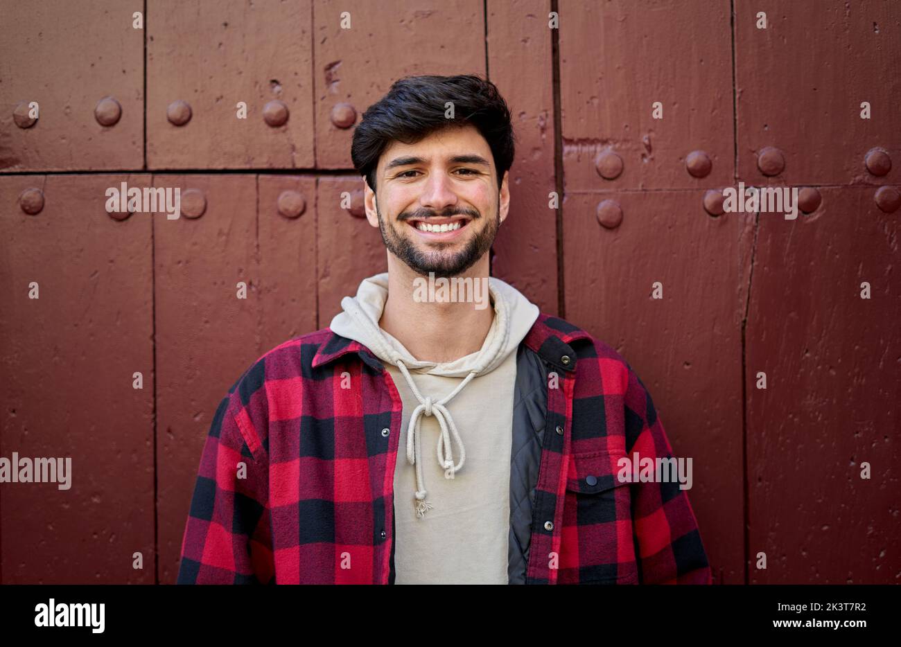 Hombre encantado con la barba mirando la cámara con una sonrisa mientras se encuentra cerca de la pared roja del edificio envejecido durante el viaje Foto de stock