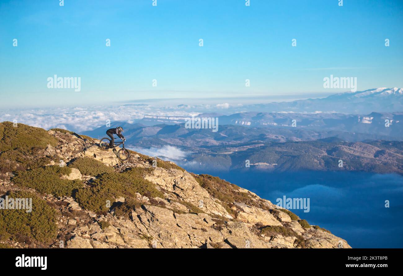 Vista lateral del chico con casco bajando en bicicleta desde la cima de la colina y pintoresca vista de las montañas en las nubes Foto de stock