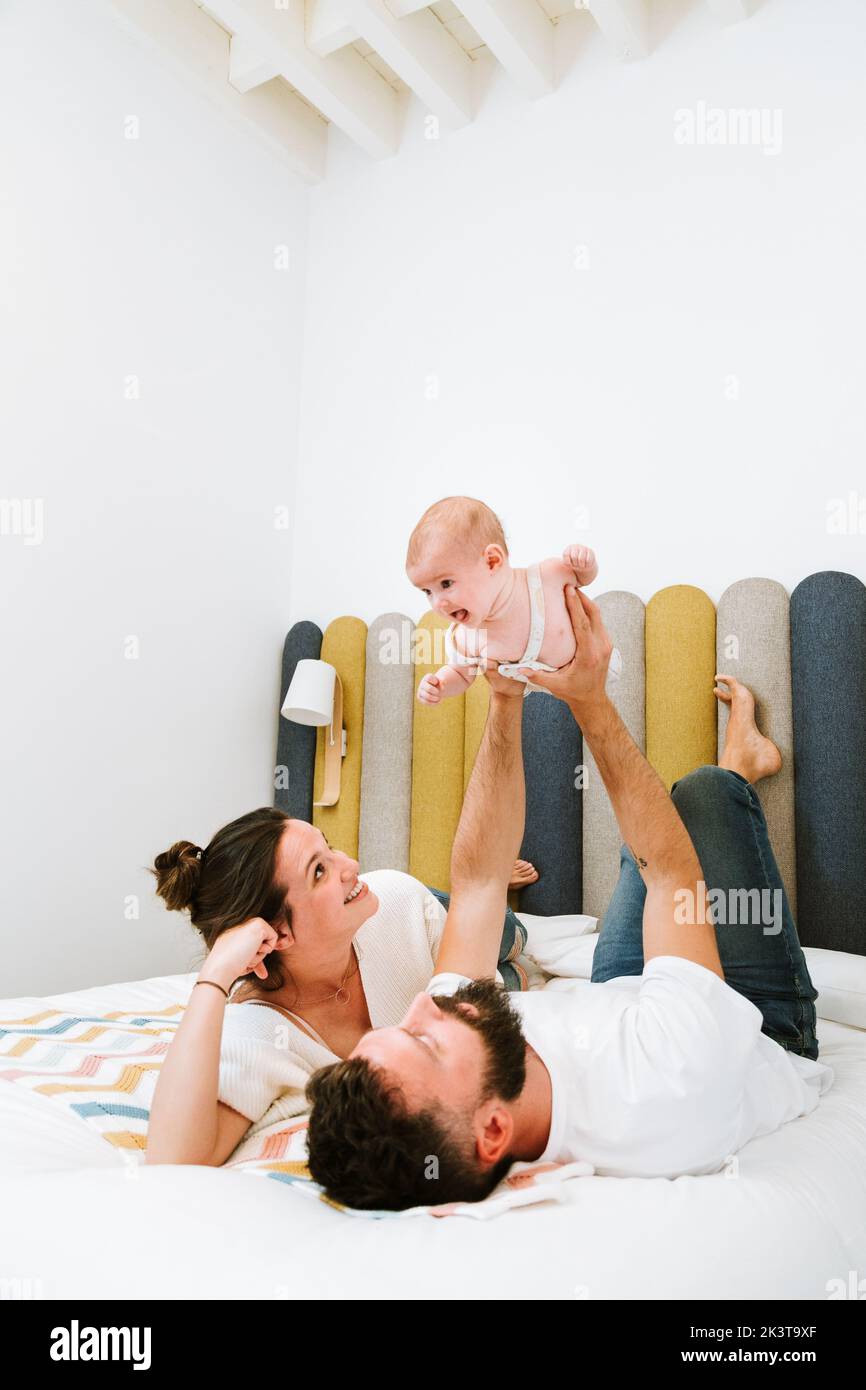 Padre fingiendo que el bebé juguetón volando como superhéroe mientras se enfría junto con la esposa y el niño en la cama suave en casa Foto de stock