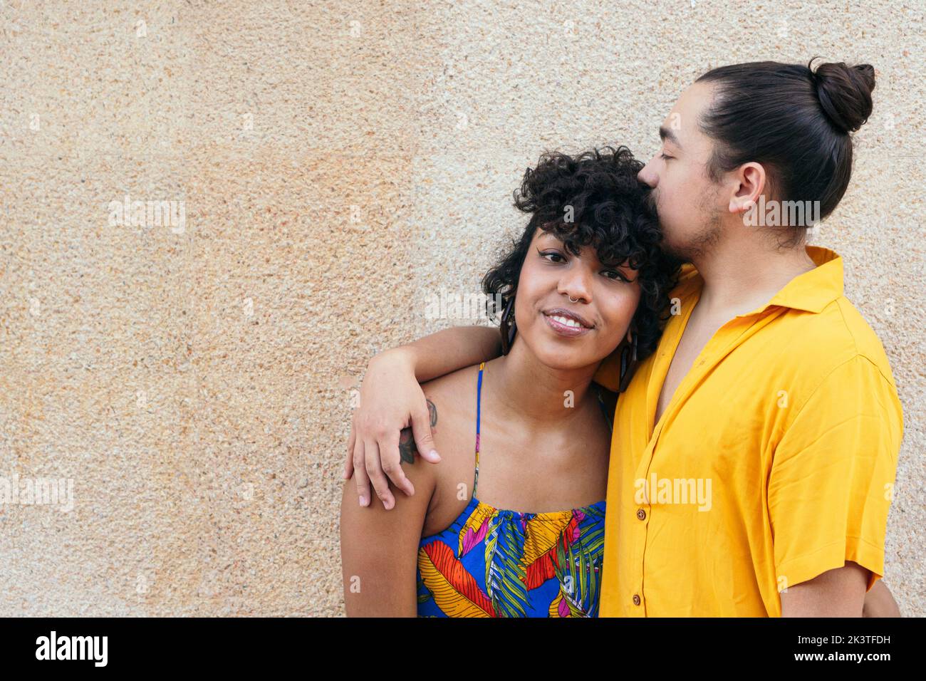 Amante hombre étnico besando novia en la cabeza mientras se encuentra contra la pared en la ciudad Foto de stock