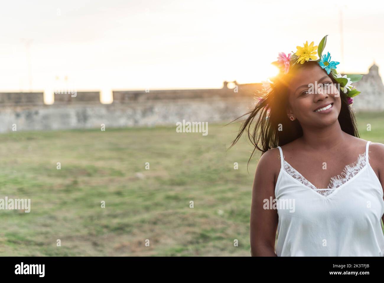 Mujer feliz con corona floral hecha de flores de colores, en el césped verde en verano y mirando la cámara Foto de stock