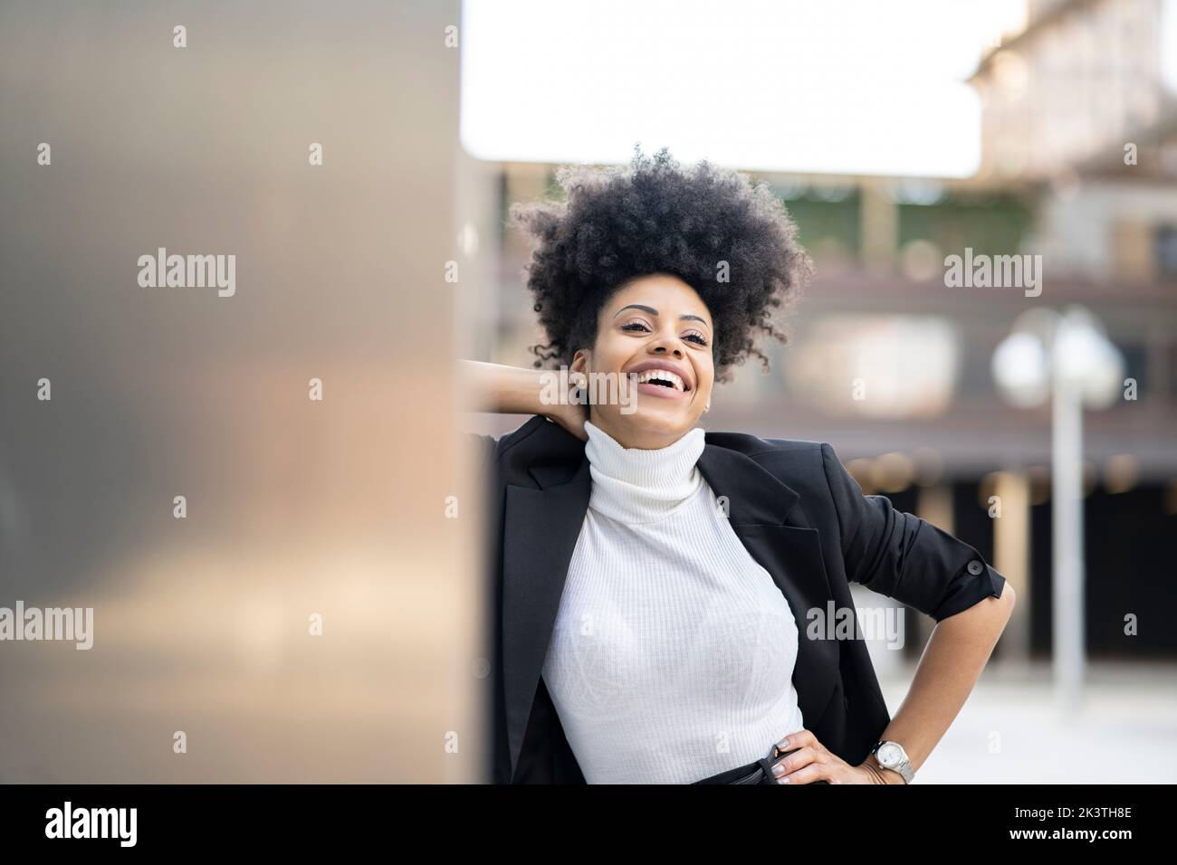 Feliz arquitecto negro en traje de moda y con peinado afro apoyado en la pared de cristal del edificio mientras que de pie mirando a distancia Foto de stock
