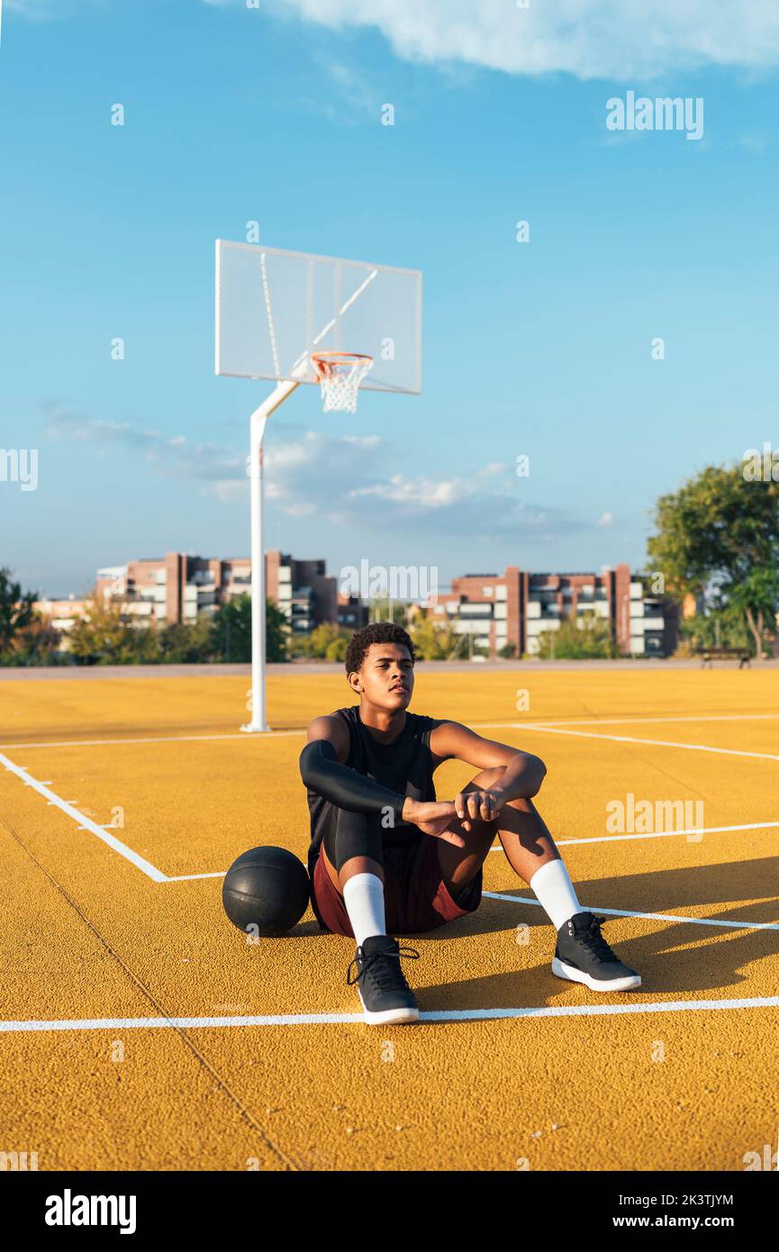Cansado deportista afroamericano en uniforme descansando sentado en el patio de recreo marcado amarillo con pelota en un día brillante Foto de stock