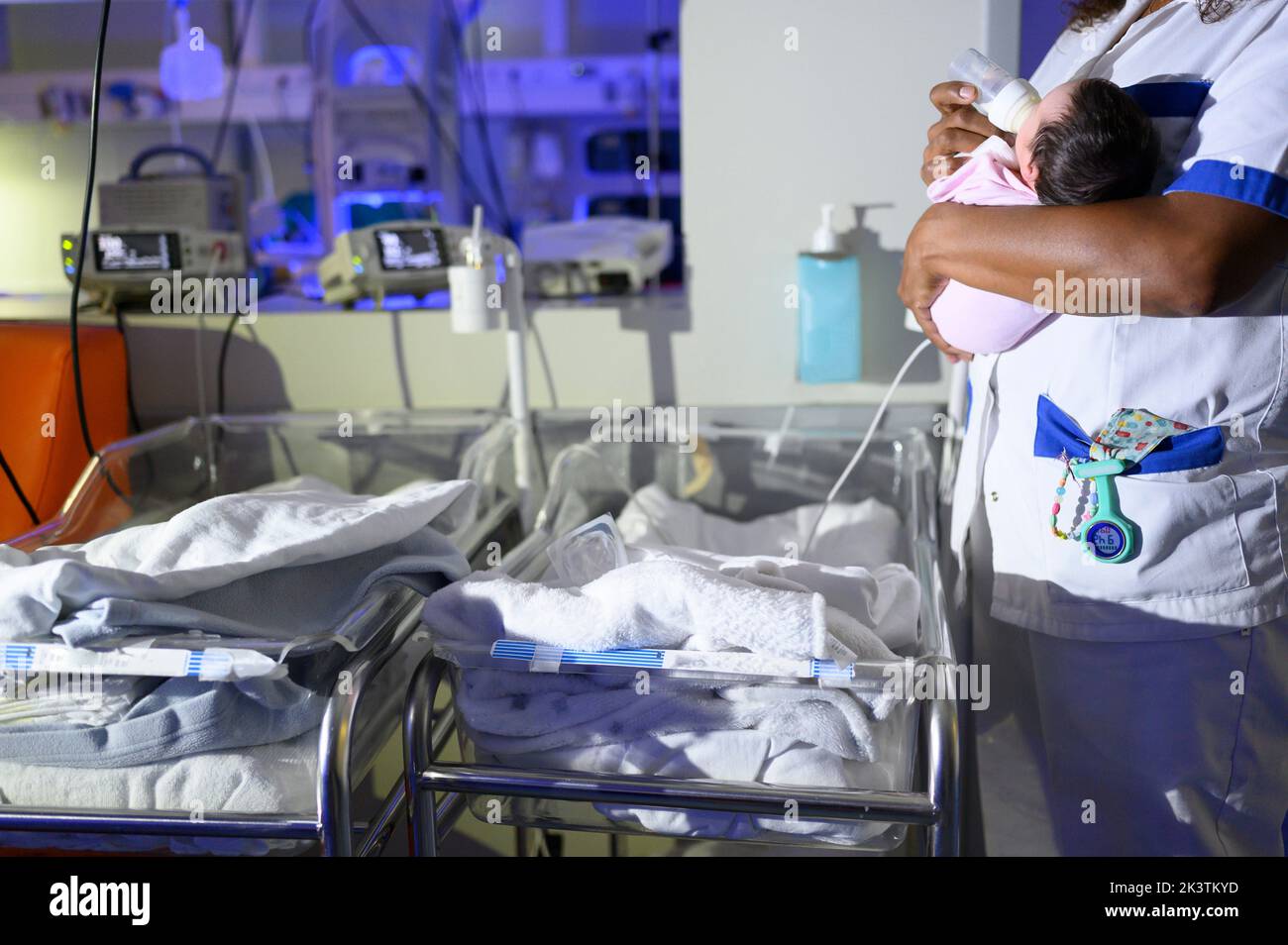 Vista lateral de la mujer de cultivo que lleva en la mano un pequeño recién nacido y la alimentación con biberón mientras está de pie junto a pequeñas camas de hospital contra el equipo médico en A. Foto de stock