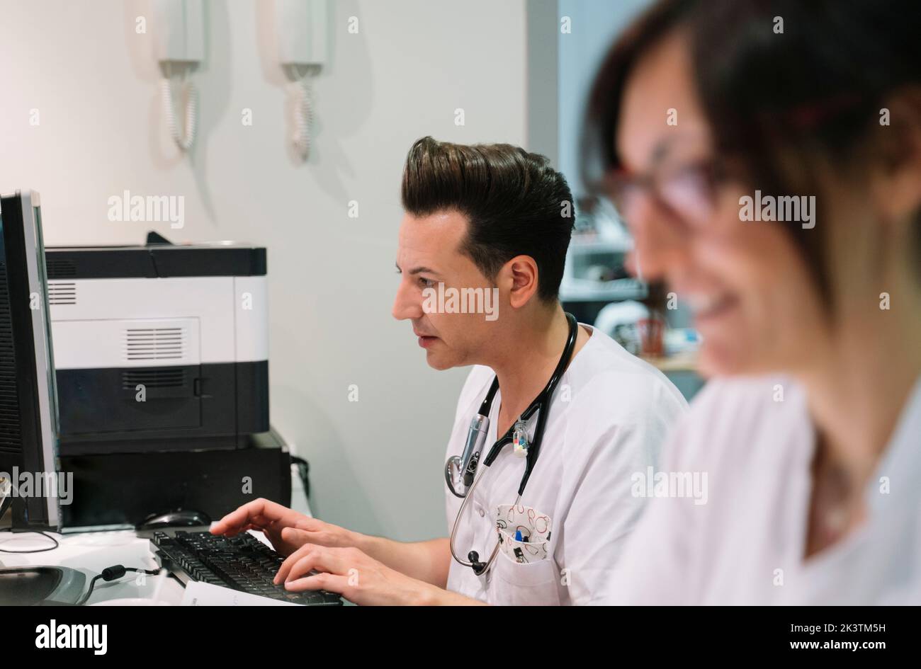 Vista lateral Doctor con enfermera anónima con uniforme blanco trabajando en la computadora en el hospital Foto de stock