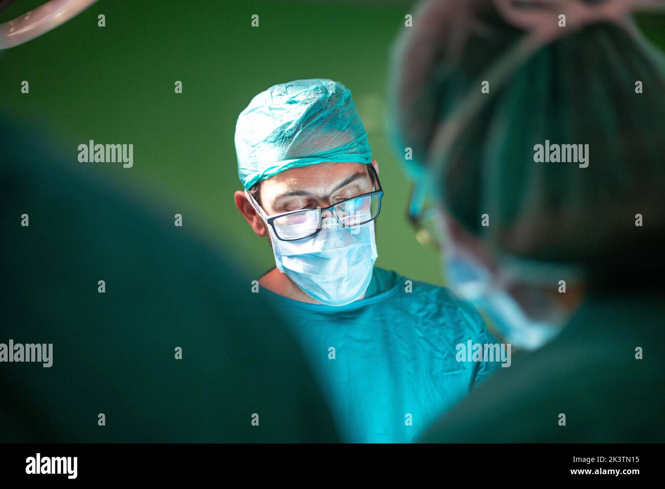 Médico varón en gafas y uniforme realizando cirugía en paciente con la ayuda de asistentes en quirófano moderno del hospital Foto de stock