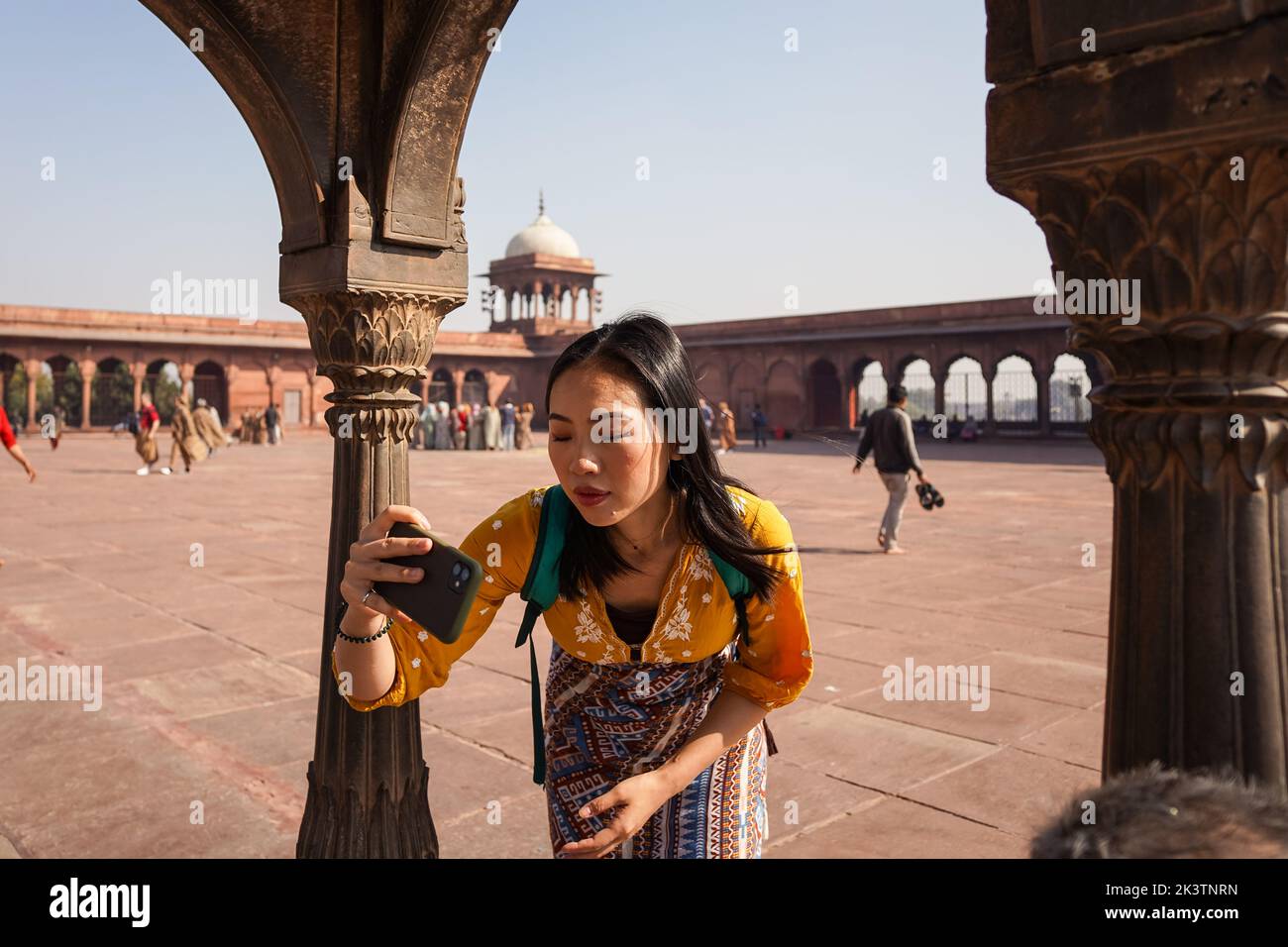 Mujer asiática en ropa ornamental con smartphone para tomar una foto mientras visita el patio de Jama Masjid en un día soleado en Nueva Delhi, India Foto de stock