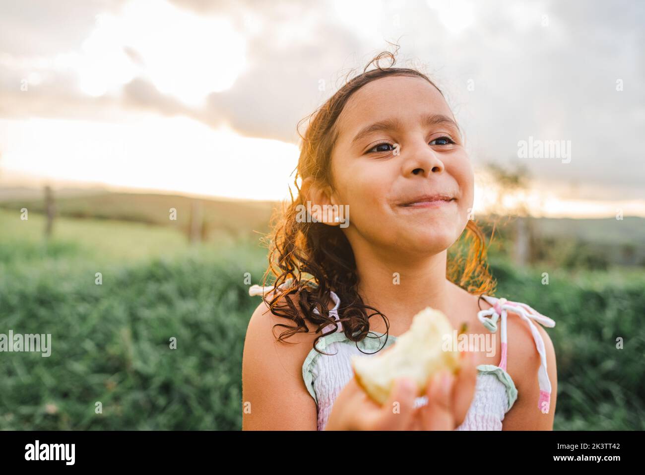 Adorable niña hispana con flor en el cabello rizado mirando lejos y morder manzana mientras descansa en la naturaleza al atardecer Foto de stock