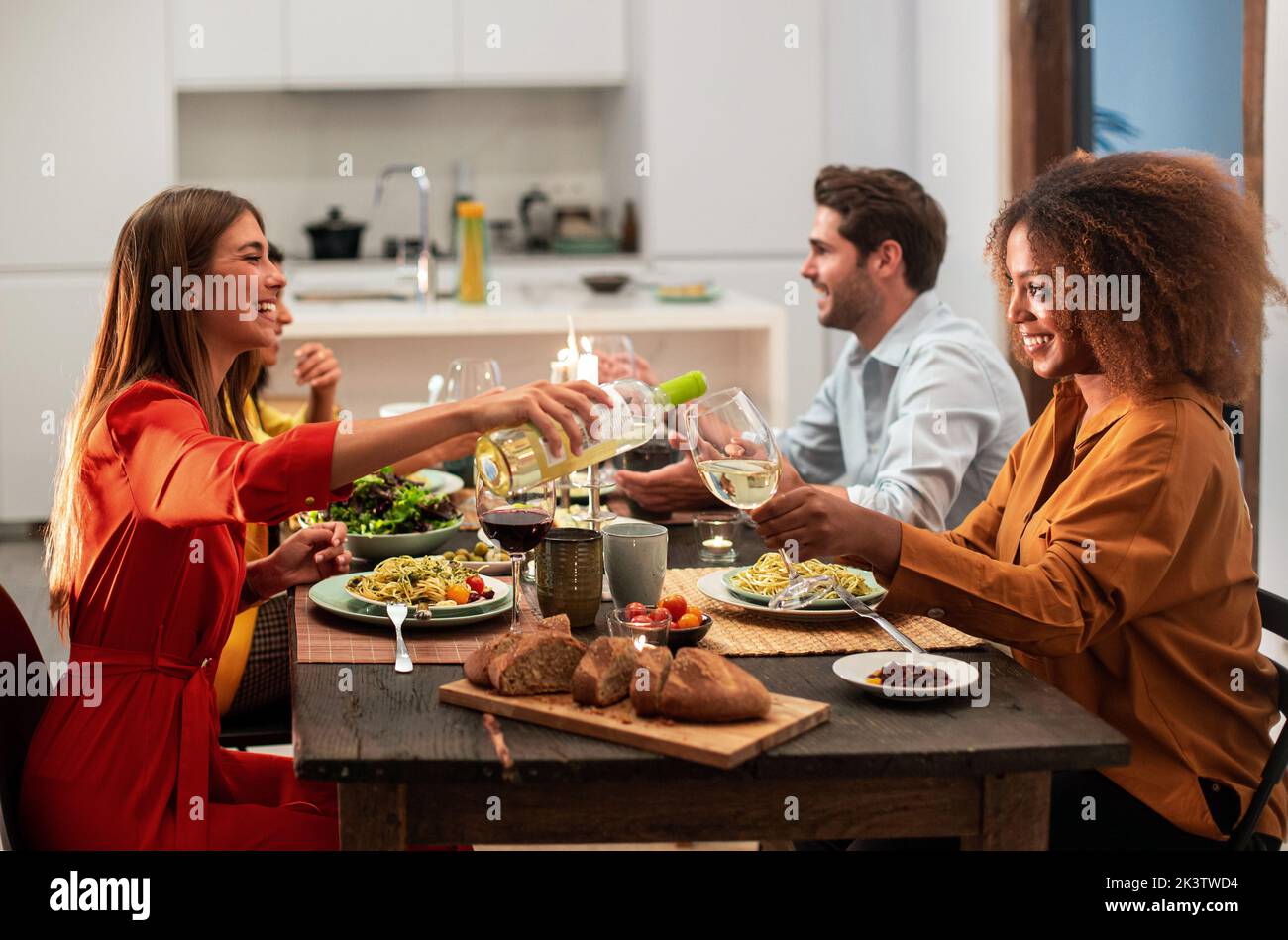Vista lateral de alegres amigos multirraciales sentados en una mesa festiva y comiendo pasta durante la celebración y fiesta en un apartamento moderno Foto de stock
