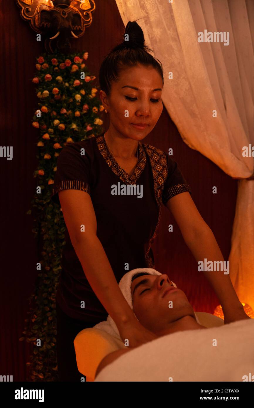 Mujer asiática con ropa tradicional que da un masaje tailandés a un hombre joven tumbado en una cama cubierta por una toalla en una habitación iluminada con luces cálidas Foto de stock