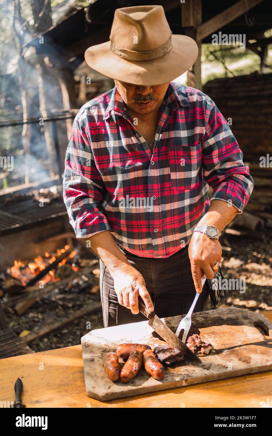 Hombre argentino en con cuchillo y tenedor cortando deliciosa pieza de carne a bordo contra el estante barbacoa a la luz del sol de stock - Alamy